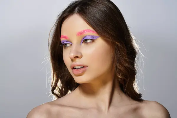 A young woman showcases bold makeup with purple eyeshadow and pink eyebrows against a grey backdrop. — Stock Photo