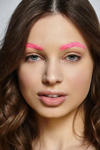 A confident young woman showcases her beauty with vibrant pink eyebrows against a grey backdrop. — Stock Photo