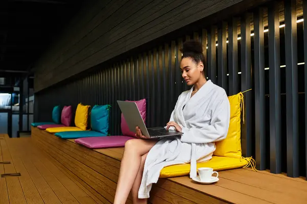 Young woman in a spa robe works on a laptop surrounded by colorful cushions, enjoying her time. — Stock Photo