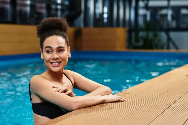 A young african american woman smiles happily while leaning at the poolside in a spa setting. — Stock Photo