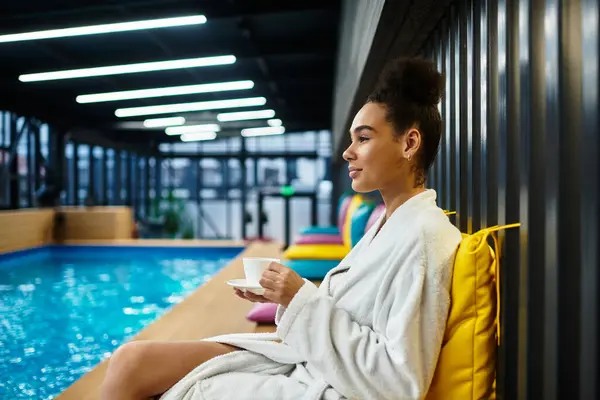 A young african american woman bathes in relaxation, sipping tea by the poolside at a spa retreat. — Stock Photo