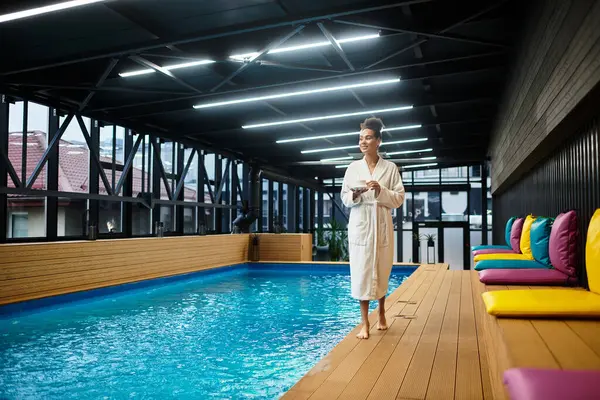 A young chic woman in a robe relaxes by a peaceful pool, embracing wellness. — Stock Photo
