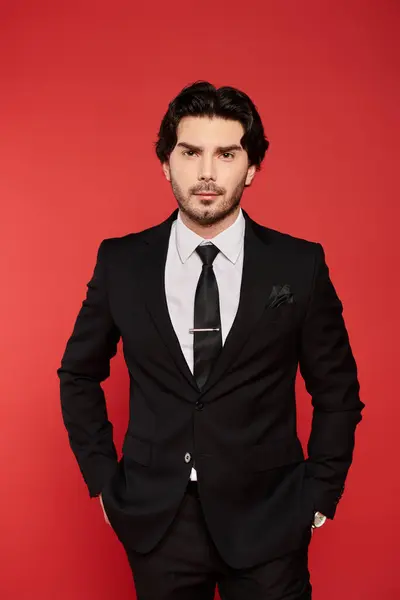 Confident young man in a black suit stands against a bold red backdrop, showcasing elegance. — Stock Photo