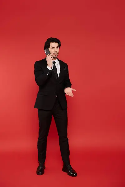 The young man stands confidently against a vibrant red backdrop, smiling as he chats on the phone. — Stock Photo