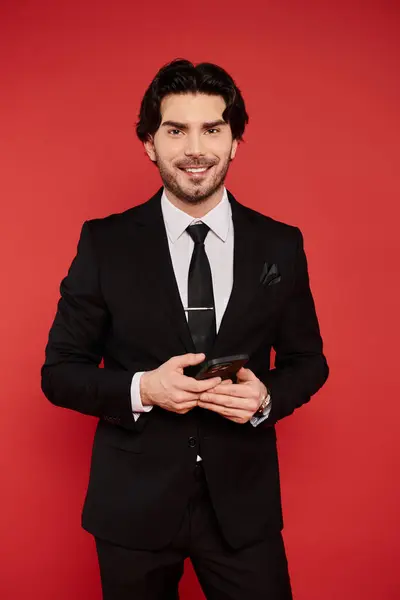 A stylish young man dressed in a black suit stands smiling, holding a smartphone, exuding charm. — Stock Photo