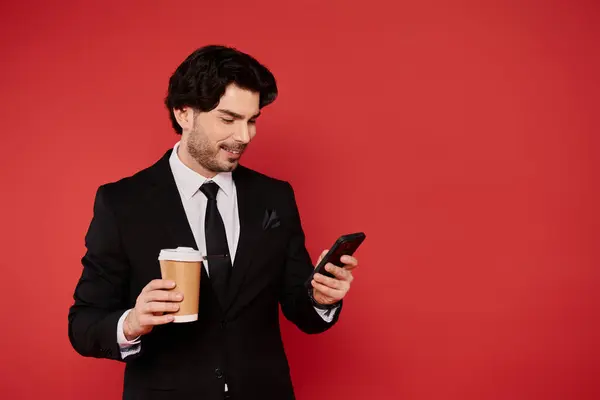 Smiling young man in a suit holds coffee cup and checks his phone in a lively setting. — Stock Photo