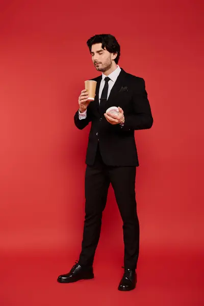 Dressed in a sleek suit, a young man savors his coffee while holding a delightful pastry. — Stock Photo