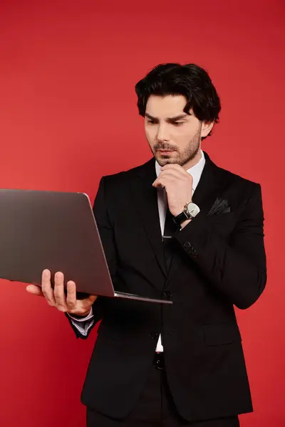 A young man in a black suit thoughtfully examines his laptop with a focused expression. — Stock Photo