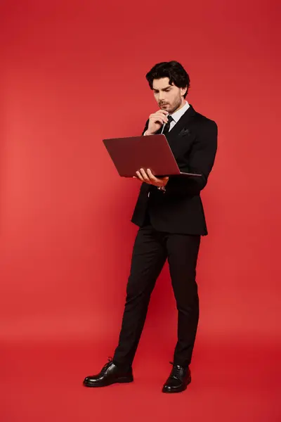 Stylish young man in formal attire thinks deeply while standing with a laptop on red background. — Stock Photo