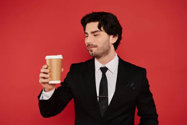 With a confident smile, the young man in a suit admires his coffee cup, radiating charm and style. — Stock Photo