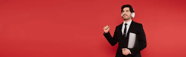 Brunette man dressed in a suit, headphones on, smiling and fist pumping in a bright red space. — Stock Photo