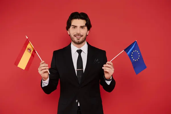 A confident young man in a sleek suit proudly holds the Spanish and EU flags. — Stock Photo