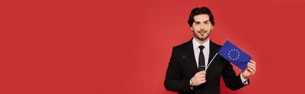 Smartly dressed man holds European Union flag, smiling broadly against a bold red backdrop. — Stock Photo