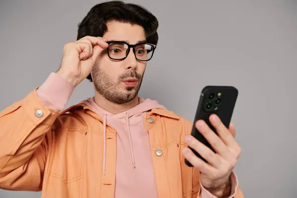 A young man with brown hair adjusts his glasses while seriously looking at his smartphone. — Stock Photo