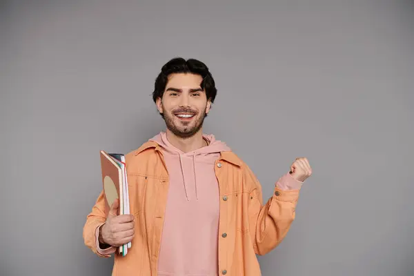 Joyful young man with brunette hair smiles brightly, holding colorful books against a gray backdrop. — Stock Photo