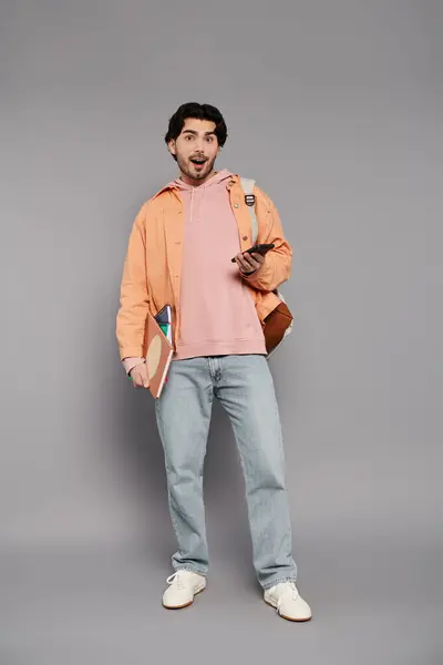 A young handsome man stands with a phone and books, showing lively enthusiasm in a studio setting. — Stock Photo