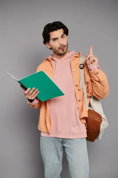 A confident young man with brown hair points while holding a green notebook. — Stock Photo