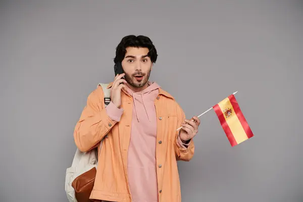 Brunette man expresses surprise while speaking on the phone and displaying a Spanish flag. — Stock Photo