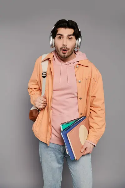 A surprised young man with headphones holds colorful notebooks in a minimalist space. — Stock Photo