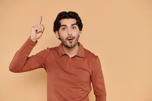 Handsome young man expresses excitement while pointing upward against a soft, warm background. — Stock Photo