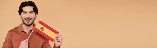 Брюнетка людина посміхається з маленьким іспанським прапором на теплому, нейтральному фоні. — стокове фото