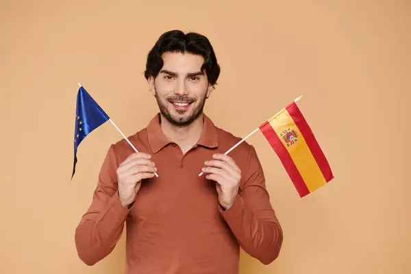 Un giovane allegro con i capelli castani regge orgogliosamente le bandiere europee e spagnole. — Foto stock