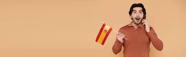 Un giovane con i capelli scuri mostra eccitazione mentre tiene in mano una bandiera spagnola e parla al telefono. — Foto stock