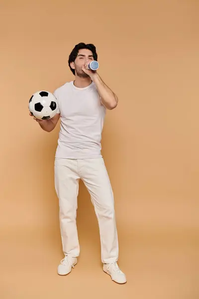 Un jeune homme aux cheveux foncés tient un ballon de football et boit dans une bouteille, rayonnant d'énergie et de joie. — Photo de stock