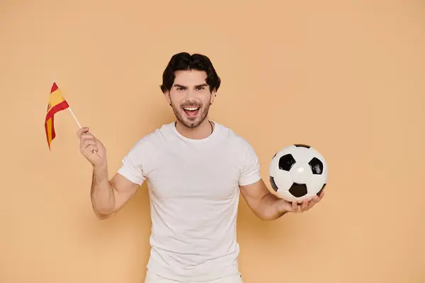 Young handsome man joyfully holds a soccer ball while waving a flag, showcasing team spirit. — Stock Photo