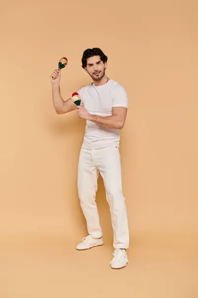 A confident young man with brunette hair joyfully plays maracas in a warm studio. — Stock Photo
