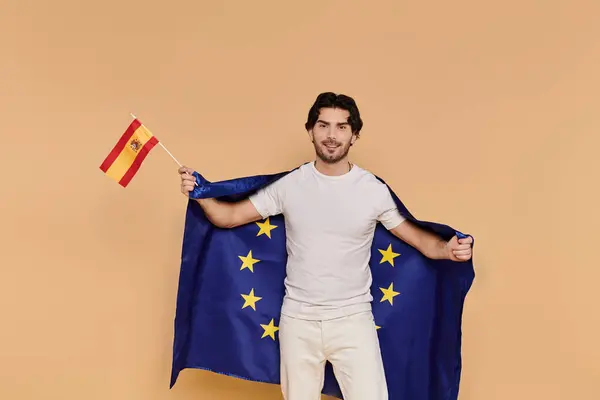 Ein hübscher junger Mann hält die spanische Flagge in die Flagge der Europäischen Union gehüllt und strahlt Freude aus. — Stockfoto
