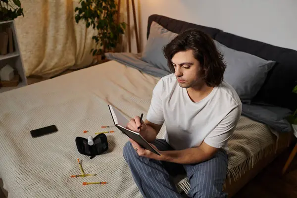 A young man with diabetes writes in his notebook while seated on his bed at home. — Stock Photo