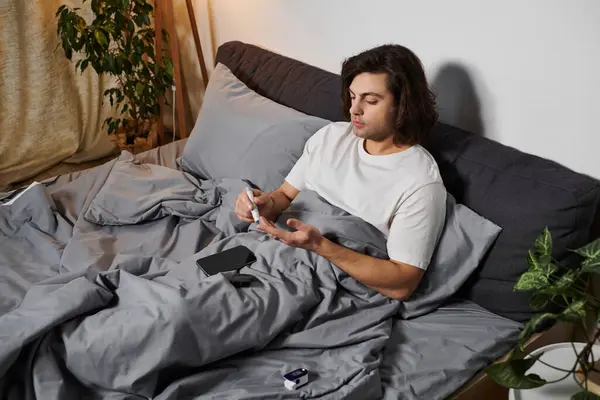 Young man checks his blood sugar levels while cozy in bed, embracing his daily routine. — Stock Photo