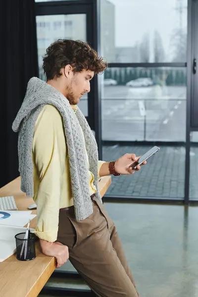 Un jeune homme aux cheveux bouclés en tenue décontractée intelligente interagit avec son téléphone au travail. — Photo de stock