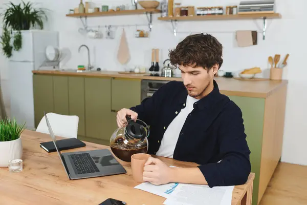Curly-haired guy is pouring coffee into a cup while focused on his work in his stylish kitchen. — Stock Photo