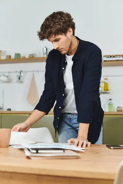 Curly-haired young man examines papers while standing at a stylish kitchen table. — Stock Photo