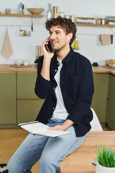 A handsome young man with curly hair chats on the phone while seated in his modern kitchen. — Stock Photo
