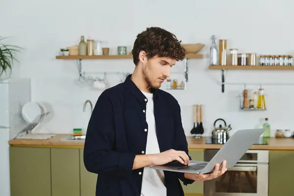 Curly-haired young man stands in his contemporary kitchen, focused on his laptop. — Stock Photo