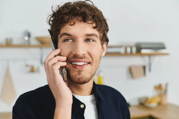 Un beau jeune homme aux cheveux bouclés sourit en parlant au téléphone dans son appartement élégant. — Photo de stock