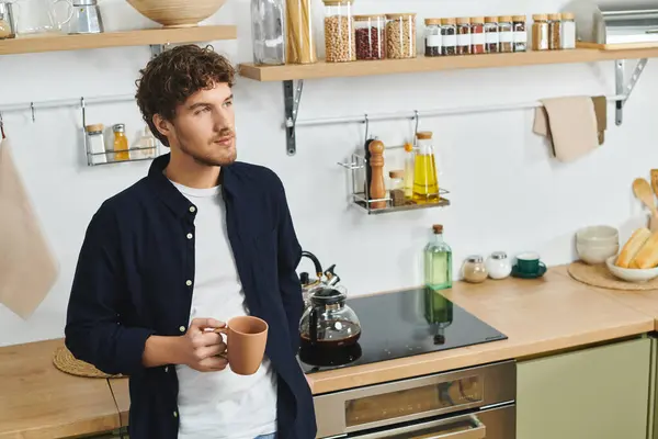 A handsome young man stands in his modern kitchen, sipping coffee and enjoying his home. — Stock Photo