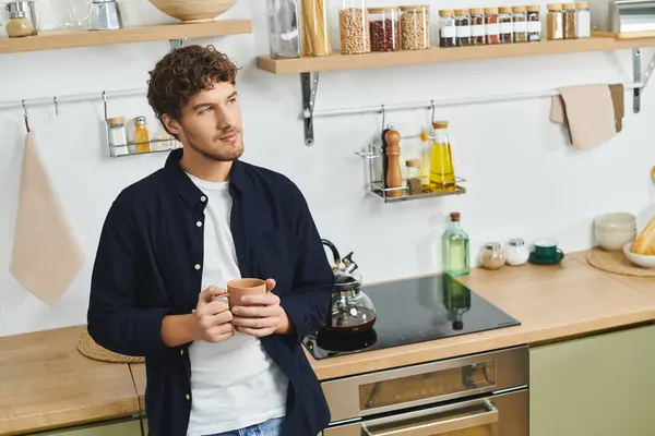 In a stylish kitchen, a young man holds a mug while gazing thoughtfully out of view. — Stock Photo