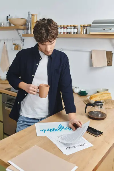 A young man with curly hair sips coffee and analyzes charts on a wooden table in his kitchen. — Stock Photo