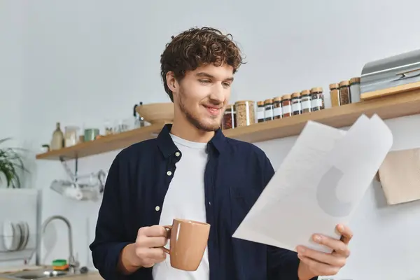 A handsome young man savoring a warm beverage as he reviews documents in his modern living space. — Stock Photo