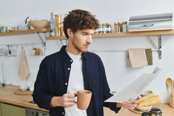 Curly-haired man in a modern kitchen reads a document while holding a warm mug. — Stock Photo