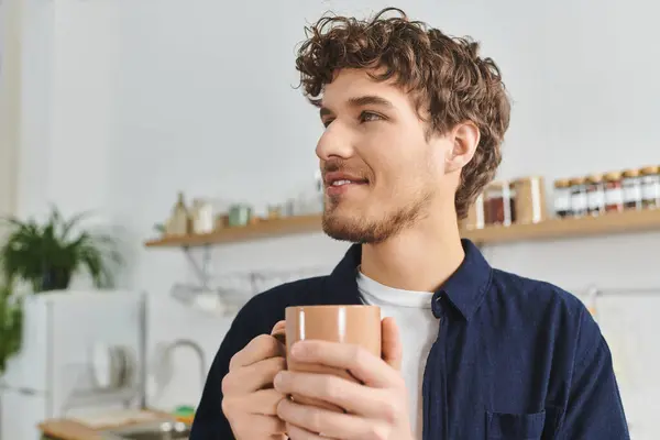 Handsome young man with curly hair smiles while holding a warm drink in his apartment. — Stock Photo