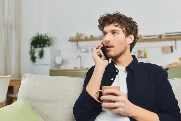 Bello giovane uomo con i capelli ricci si rilassa a casa, tenendo una tazza e chiacchierando al telefono. — Foto stock