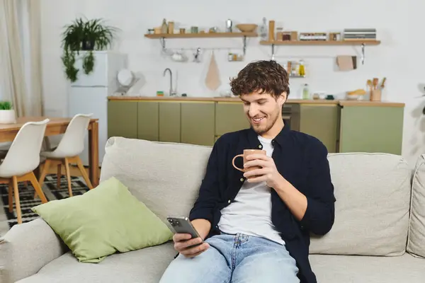 Un joven con el pelo rizado se sienta en un sofá, bebiendo café y mirando su teléfono. - foto de stock