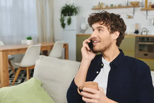 A young curly-haired man laughs and chats on the phone in his stylish apartment. — Stock Photo