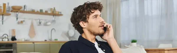 A young man with curly hair relaxes in a stylish apartment while talking on his phone, banner — Stock Photo