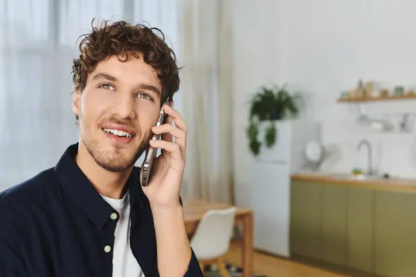 Curly-haired young man smiles while talking on a phone in his modern, cozy apartment. — Stock Photo